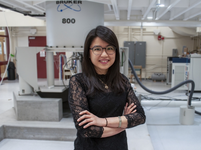 A woman with glasses and arms crossed stands smiling in front of a large Bruker 800 NMR instrument in a research lab.