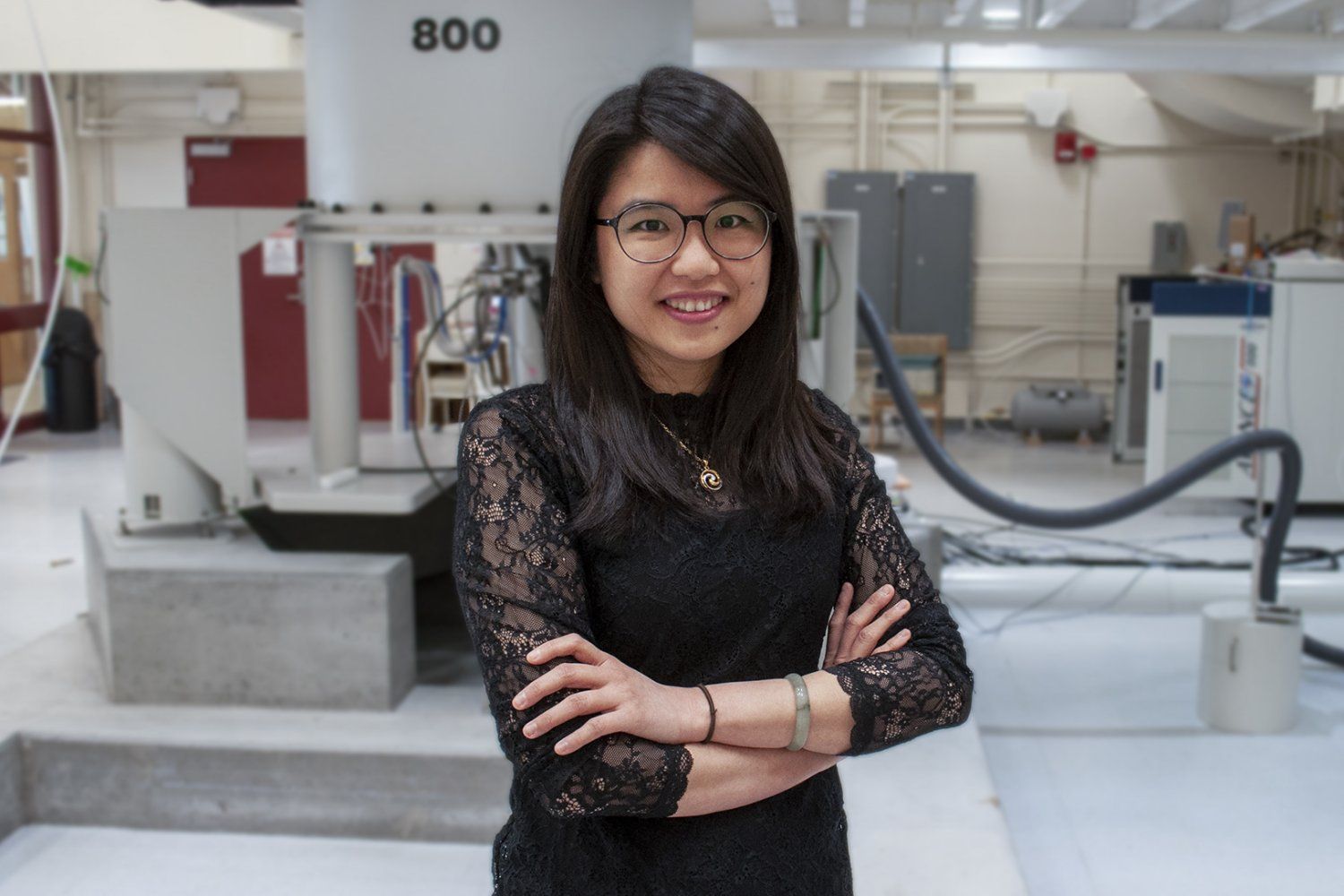 A woman with glasses and arms crossed stands smiling in front of a large Bruker 800 NMR instrument in a research lab.