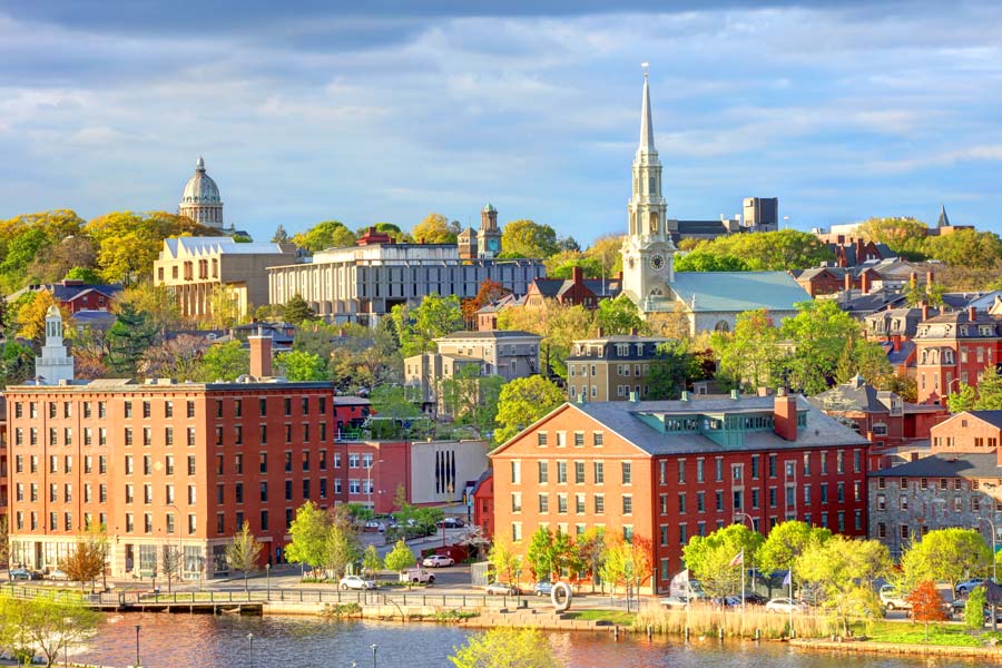 Providence, Rhode Island waterfront skyline in spring, with historic brick mill buildings along the river, a tall white church steeple, and the State House dome visible on the hill.
