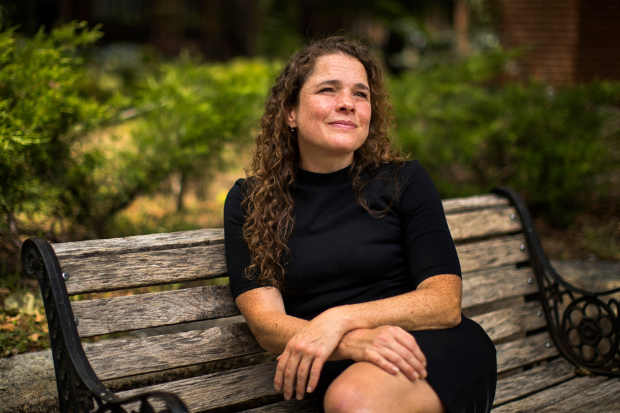MIT researcher Sarah Williams sits on a wooden park bench outdoors, looking upward thoughtfully, wearing a black dress with lush green garden shrubs visible behind her.