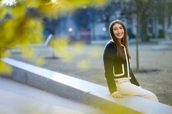 MIT PhD candidate Shaylin Cetegen sits on a stone wall with foliage in the foreground.