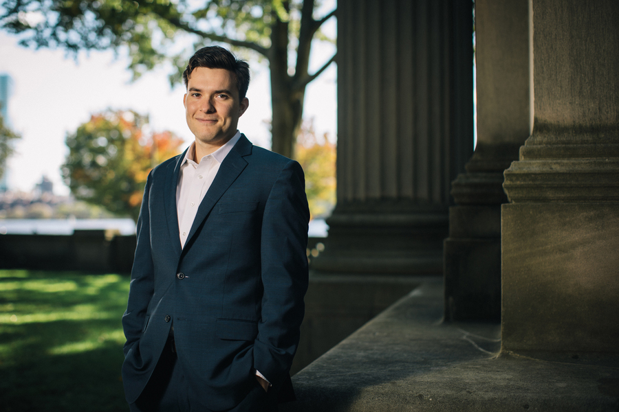 Portrait of an MIT researcher in a navy blazer leaning against a stone column on campus, with the Charles River and fall foliage visible in the background.