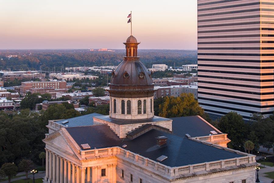 Close aerial view of the South Carolina State House dome in Columbia at dusk, its copper roof glowing in warm light with the city stretching to the tree-lined horizon.