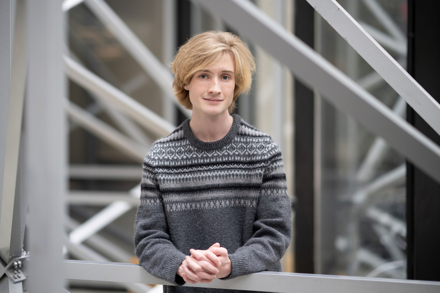A young MIT student with blond hair and a gray Fair Isle sweater stands with hands clasped in front of a steel staircase structure inside an MIT research building