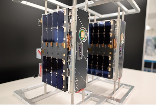 On a tabletop, two small, boxy satellites sit, roughly 20 x 10 x 30 cm in size. 