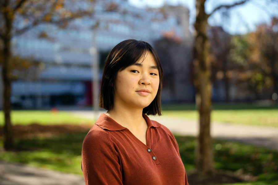 An MIT student poses outdoors on a tree-lined campus path wearing a rust-colored polo shirt, with a glass building and green lawn visible in the soft-focus background.