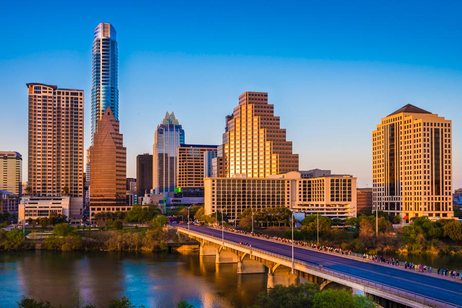 Austin, Texas skyline bathed in golden sunset light along Lady Bird Lake, with pedestrians crossing the Congress Avenue Bridge in the foreground.
