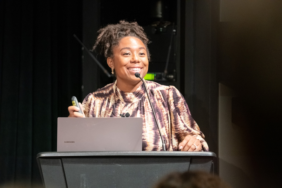 An MIT researcher smiling at a podium with a laptop and microphone while presenting at a lecture or conference.