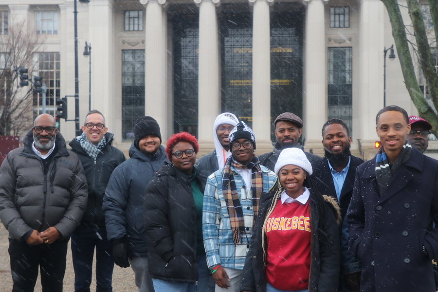 Group of approximately 10 diverse people in winter clothing pose in snow before classical building with white columns.