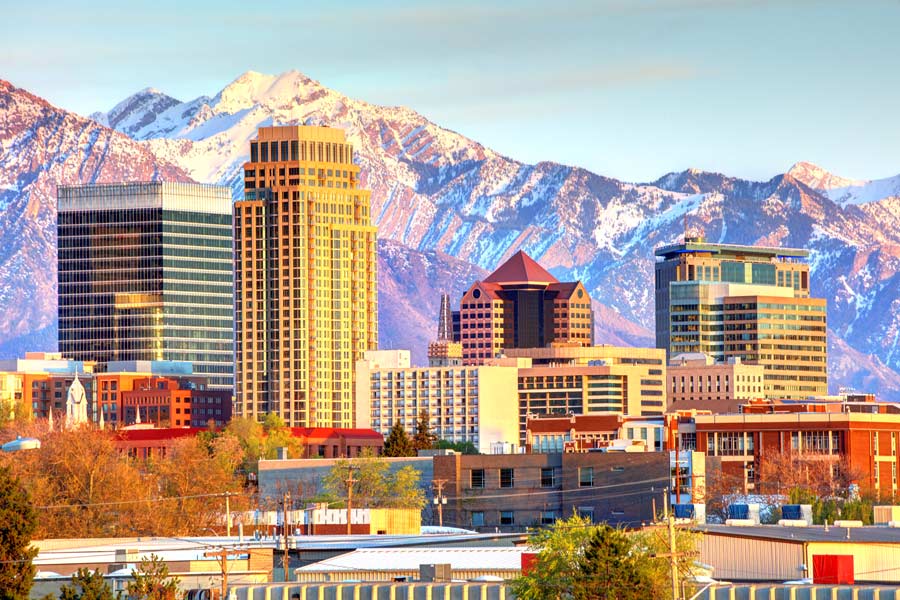 Salt Lake City, Utah skyline bathed in warm light with snow-capped Wasatch Mountains rising dramatically behind downtown skyscrapers.