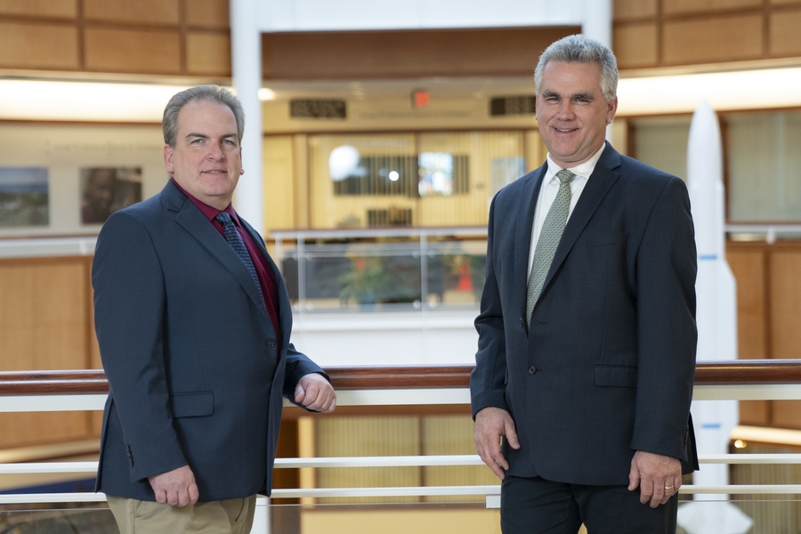 Two men in dark business suits and ties stand together in a modern institutional hallway with wood paneling, glass windows, and metal railings in the background.