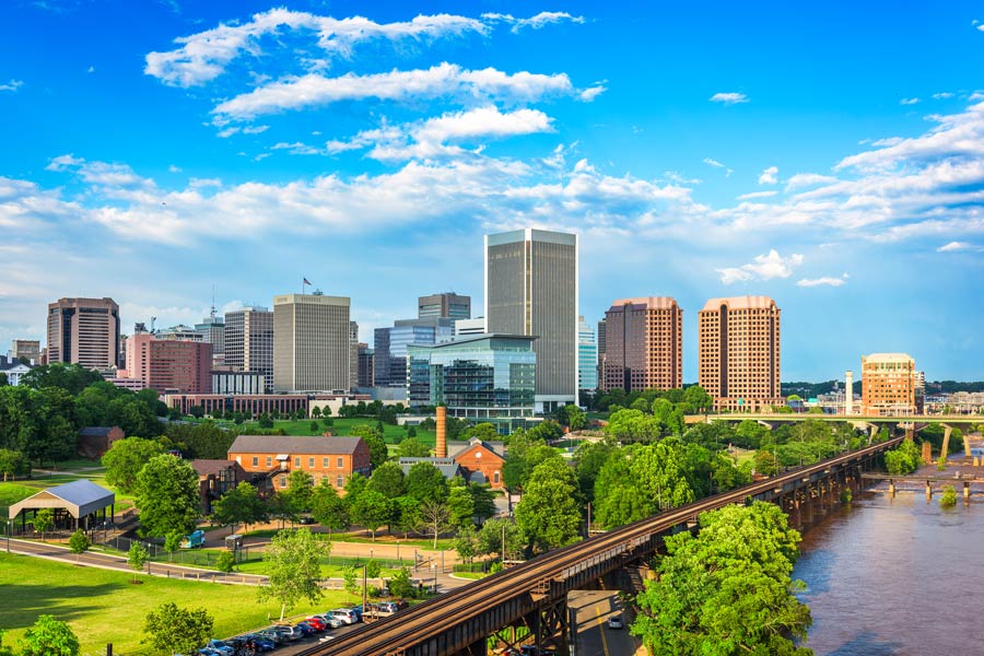 Richmond, Virginia skyline along the James River, with a railroad bridge and historic Tredegar Iron Works buildings in the foreground surrounded by lush summer greenery.