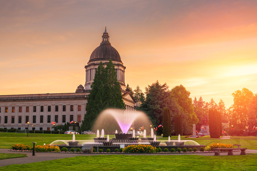The Washington State Capitol building in Olympia at sunset, with an illuminated fountain and flower gardens in the foreground.