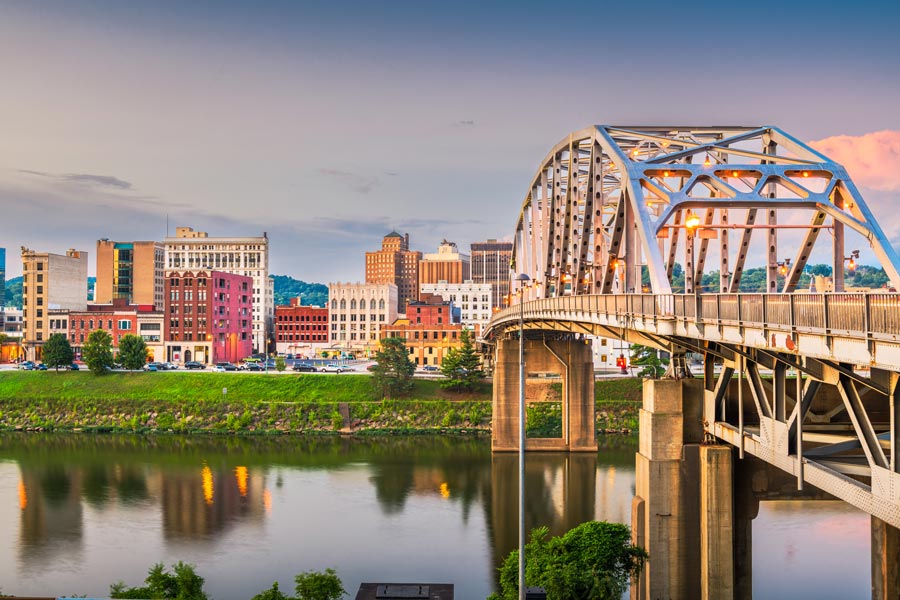 Charleston, West Virginia skyline at dusk reflected in the Kanawha River, with a steel arch bridge prominently spanning the foreground.