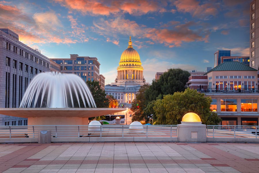 The illuminated Wisconsin State Capitol dome at sunset, viewed from Monona Terrace with a large fountain in the foreground.