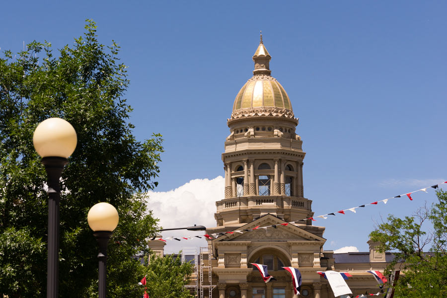 The Wyoming State Capitol building in Cheyenne, with its ornate gold dome decorated with bunting, framed by summer trees and globe street lamps against a blue sky.