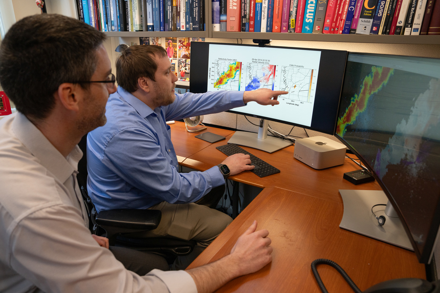 Two men at a desk review colorful weather radar and probability maps displayed across two large monitors, with a bookshelf full of textbooks behind them.