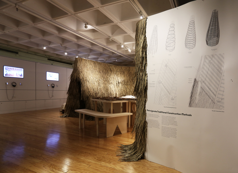 Gallery installation featuring a large thatched structure made of bundled natural fibers beside architectural drawings and diagrams exploring traditional construction methods.