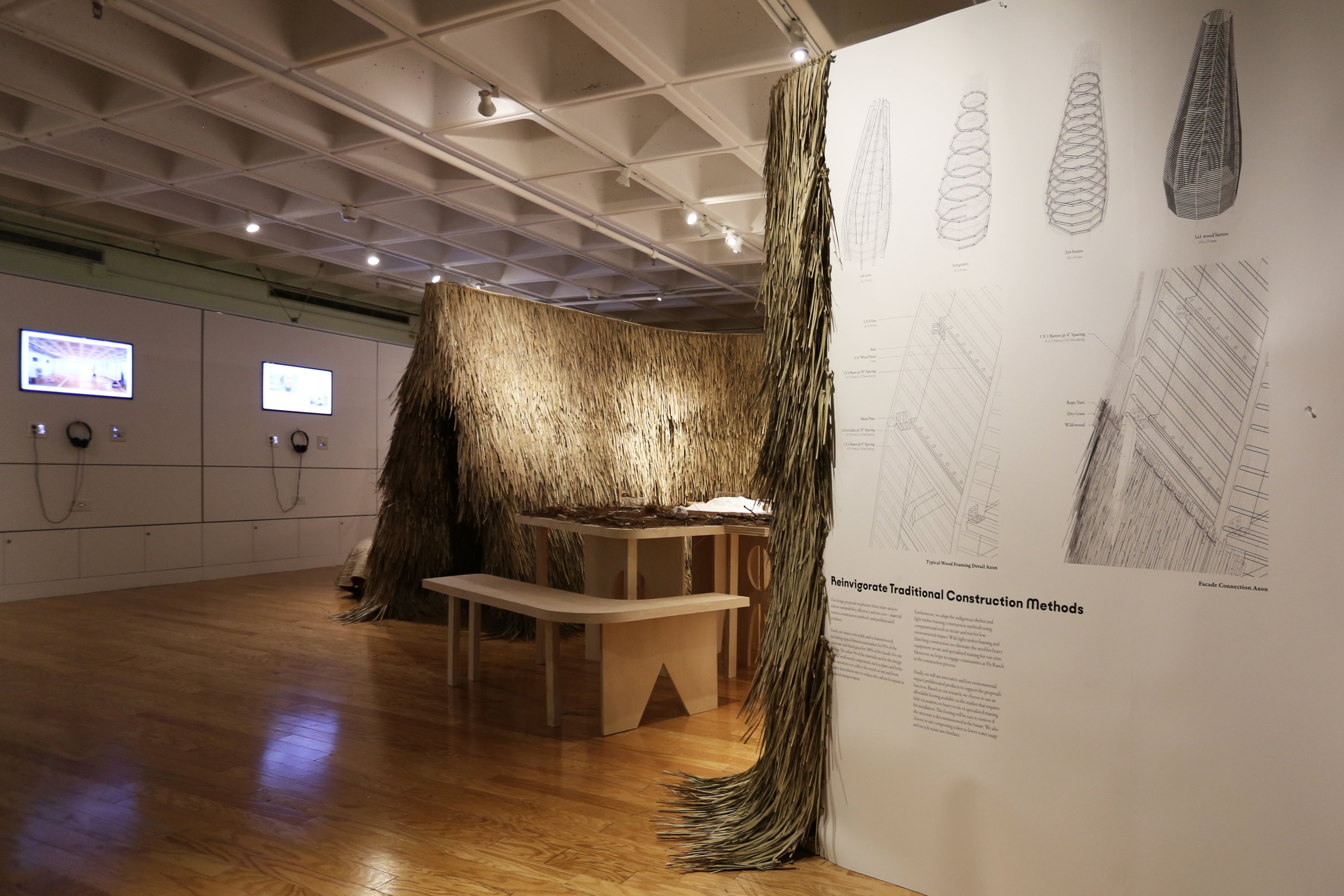 Gallery installation featuring a large thatched structure made of bundled natural fibers beside architectural drawings and diagrams exploring traditional construction methods.