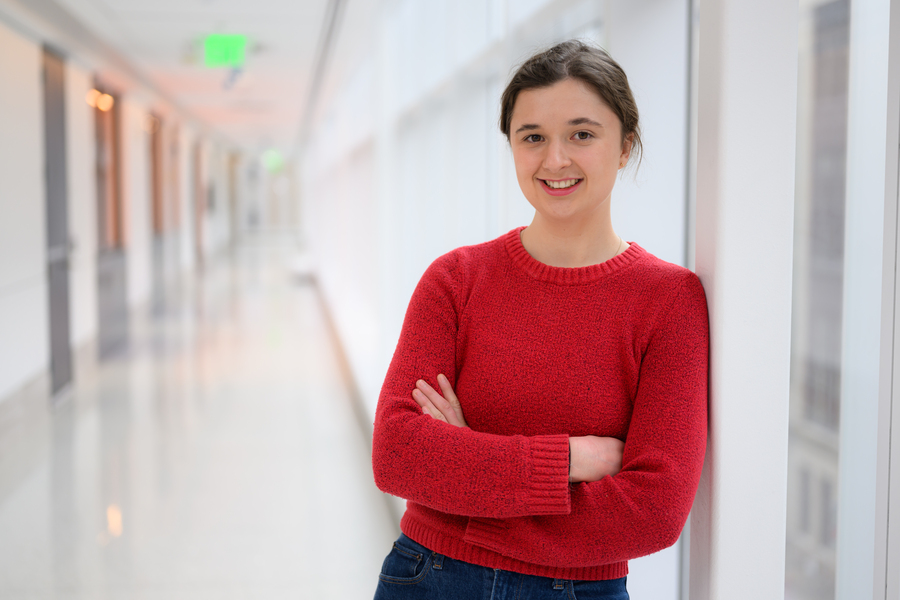 A smiling young woman in a red sweater stands with arms crossed in a bright modern corridor.