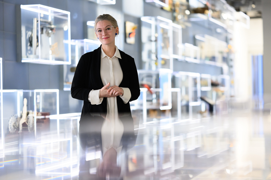 A woman in a black blazer and white blouse stands with hands clasped in a modern museum gallery lined with illuminated display cases.