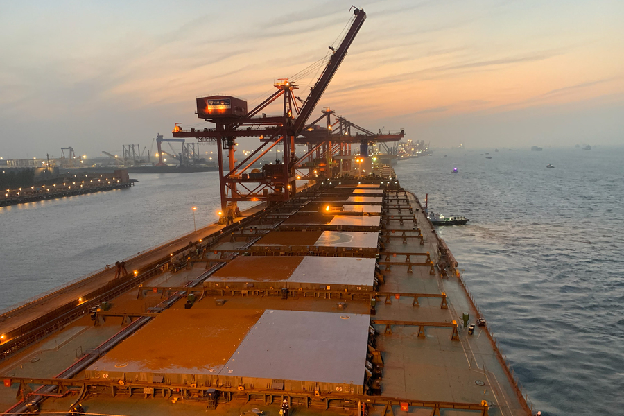 Aerial view along the deck of a large bulk carrier cargo ship at dusk, with port cranes and harbor lights illuminating the waterway,