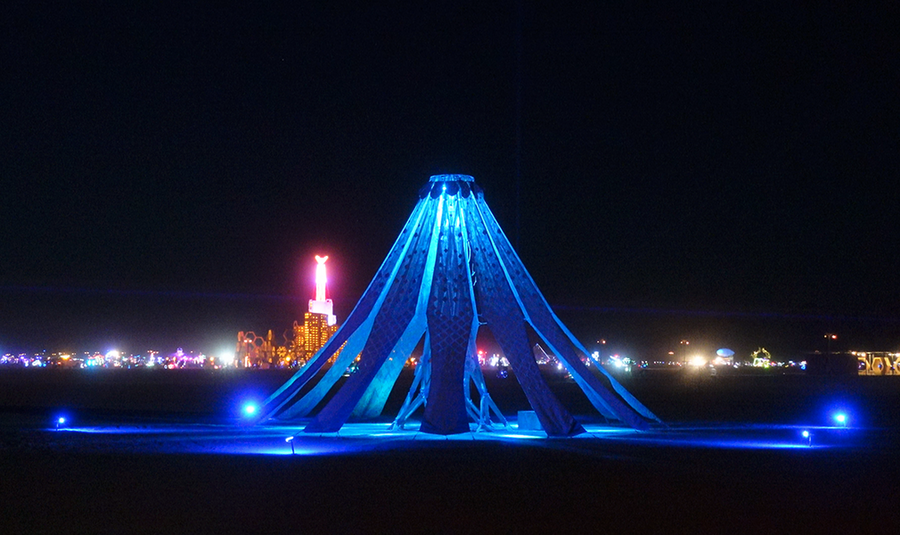 A large wooden sculptural installation lit in blue at night on an open desert playa, with the glowing lights of Burning Man visible on the horizon.