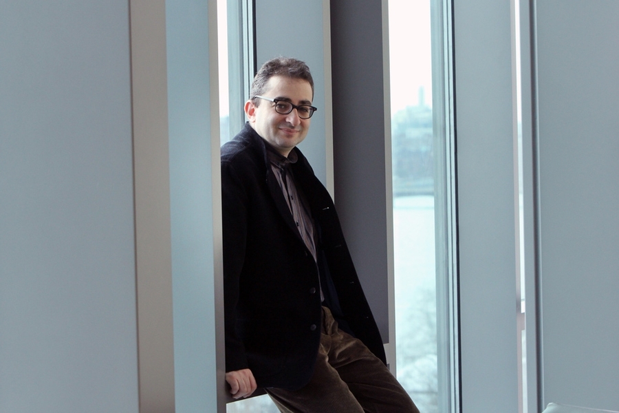 A man in a dark blazer and glasses leans against a wall between two windows, smiling over his shoulder at the camera.