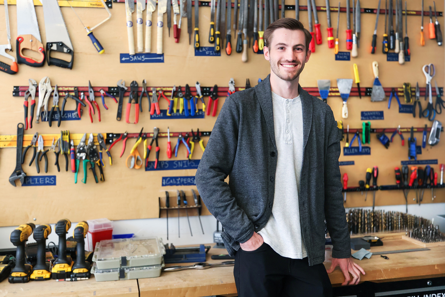 A smiling MIT fellow stands in front of a pegboard wall organized with labeled rows of hand tools including saws, pliers, and calipers, with power drills and drill bits on the workbench beside him.