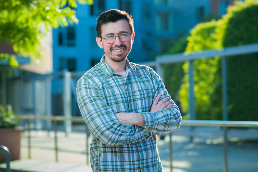 Portrait of an MIT researcher in a green plaid shirt with arms crossed, smiling outdoors on a sunny campus terrace.