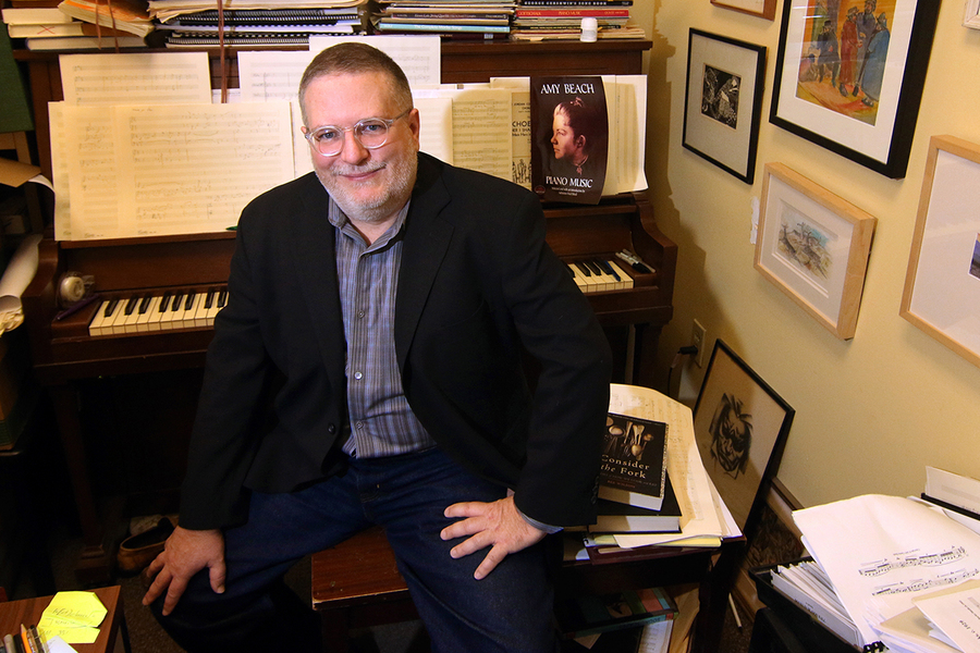 A man in a black blazer and glasses sits at a piano surrounded by sheet music, framed artwork, and open books including a volume of Amy Beach piano music.
