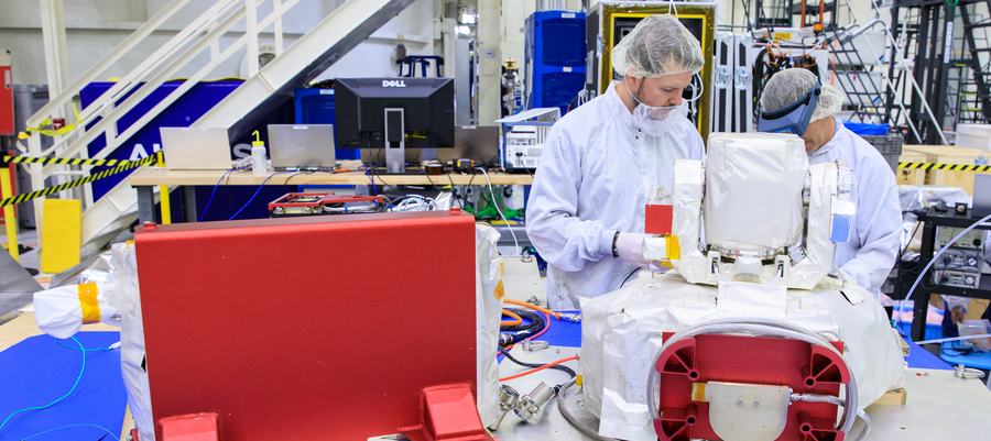 Two MIT Lincoln Laboratory technicians in white cleanroom suits and hairnets assemble a communications terminal component in a large research facility, with red hardware enclosures and lab workstations visible in the background.