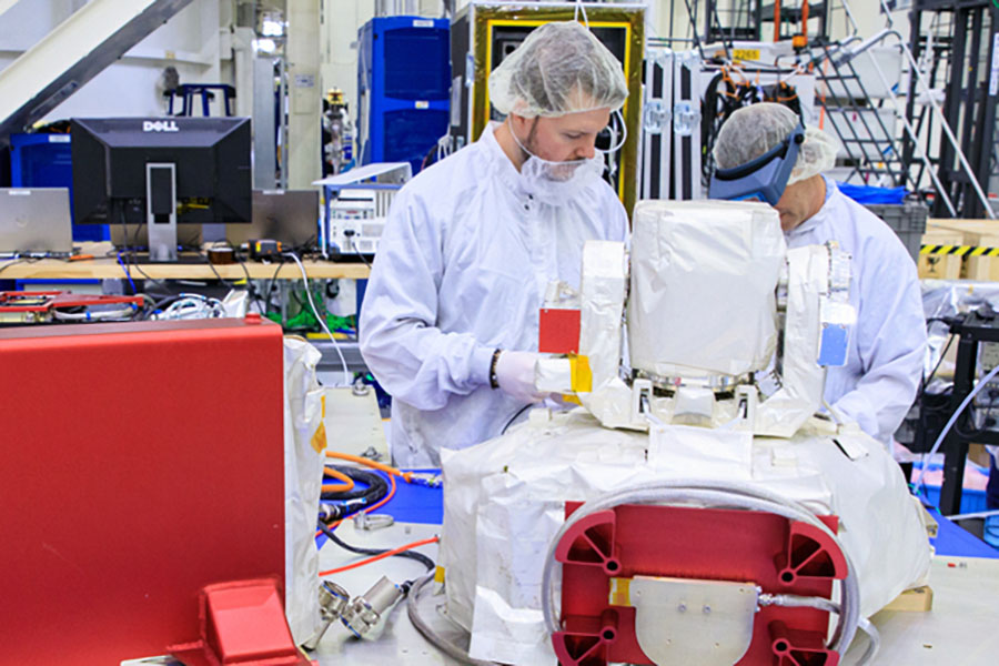 Two MIT Lincoln Laboratory technicians in white cleanroom suits and hairnets assemble a communications terminal component in a large research facility, with red hardware enclosures and lab workstations visible in the background.