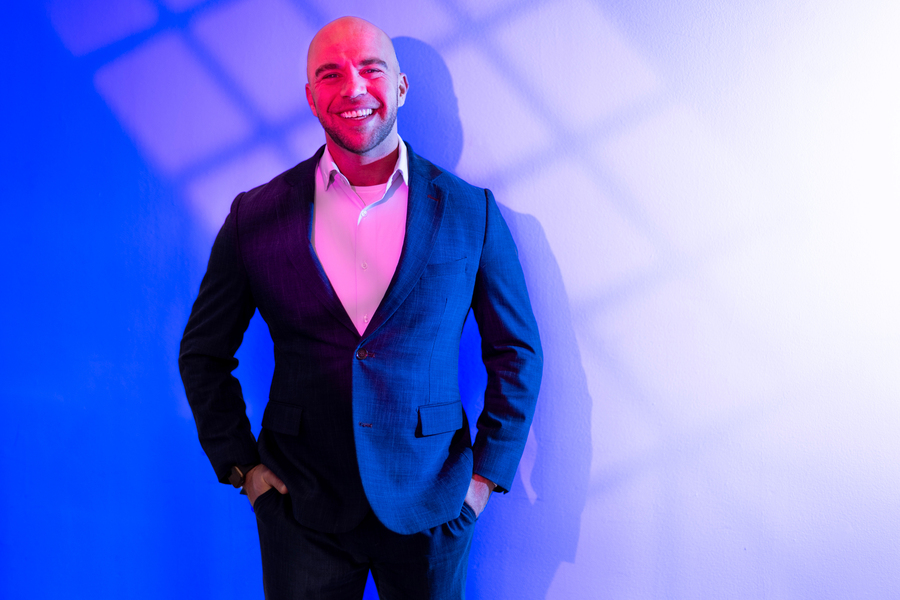 A man in a navy suit smiles confidently against a blue and white geometric light projection background, with dramatic red and blue studio lighting creating a bold shadow effect.