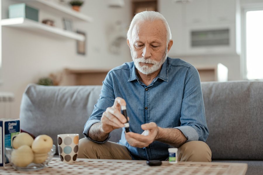 An older man with white hair and a beard uses a blood glucose monitor while sitting on a couch at home, with a bowl of fruit and a Contour glucometer box visible on the coffee table in front of him.