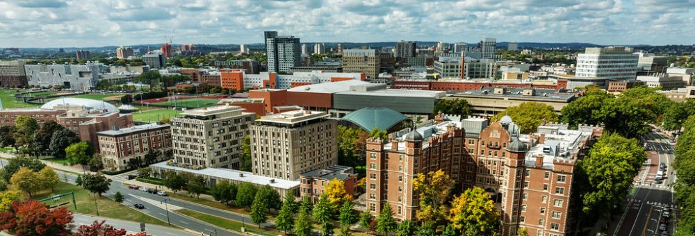 Aerial view of the MIT campus with academic buildings, sports fields, trees, and a city skyline in the background under a partly cloudy sky.