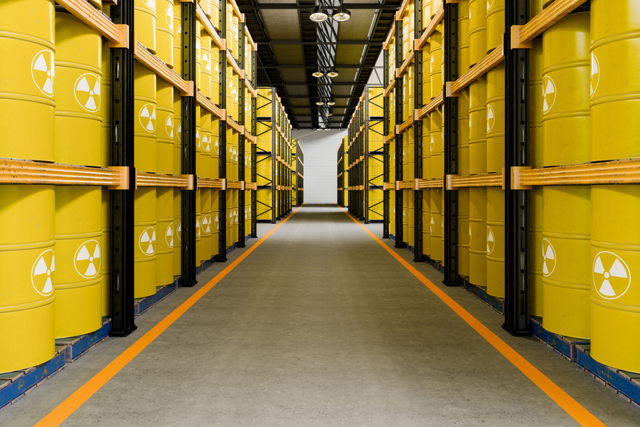 Long warehouse aisle lined floor-to-ceiling with yellow barrels marked with radioactive hazard symbols on industrial shelving.