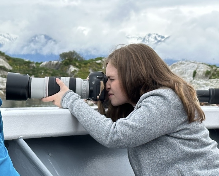 A researcher leans over the railing of a boat and aims a large telephoto camera lens toward the water, with snow-capped mountains and rocky coastline visible in the background.
