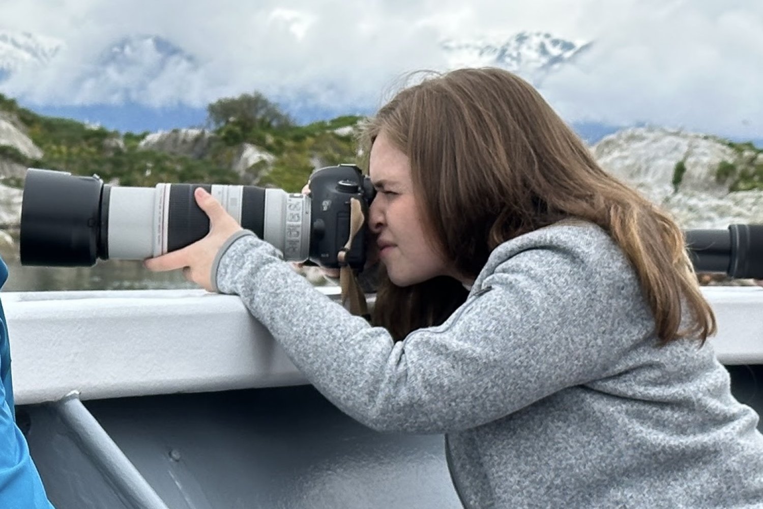 A researcher leans over the railing of a boat and aims a large telephoto camera lens toward the water, with snow-capped mountains and rocky coastline visible in the background.