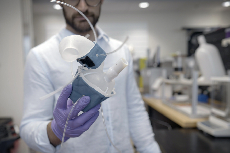 A researcher in a white lab coat and purple gloves holds a blue 3D-printed heart model with attached tubing in a laboratory setting.