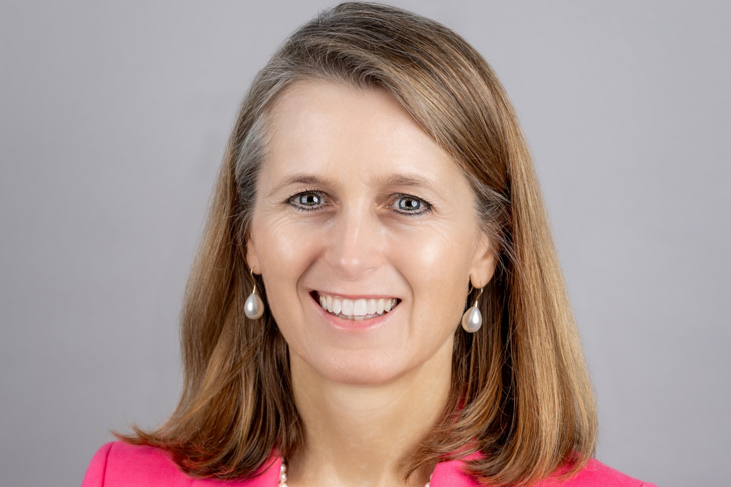 A headshot of a woman in a pink blazer with pearl jewelry, smiling against a gray background.