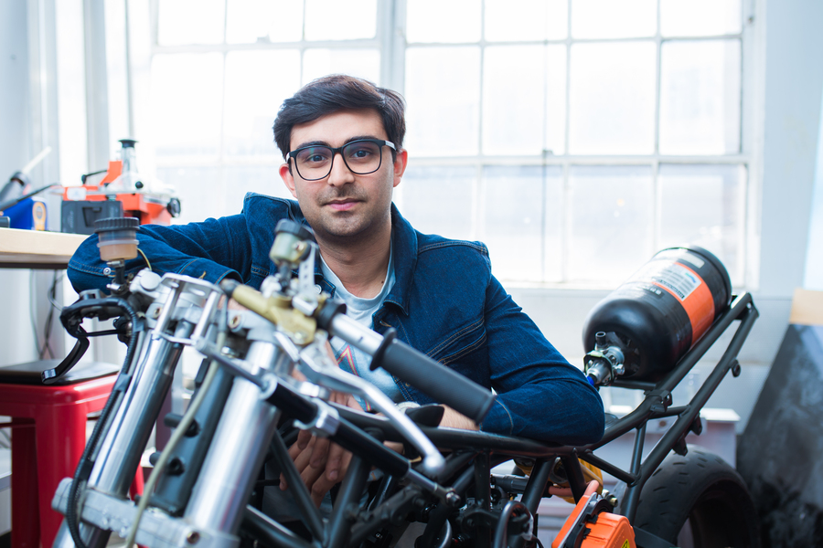 A researcher in glasses and a denim jacket leans over a motorcycle chassis with exposed engine components and a pressurized tank in a bright workshop