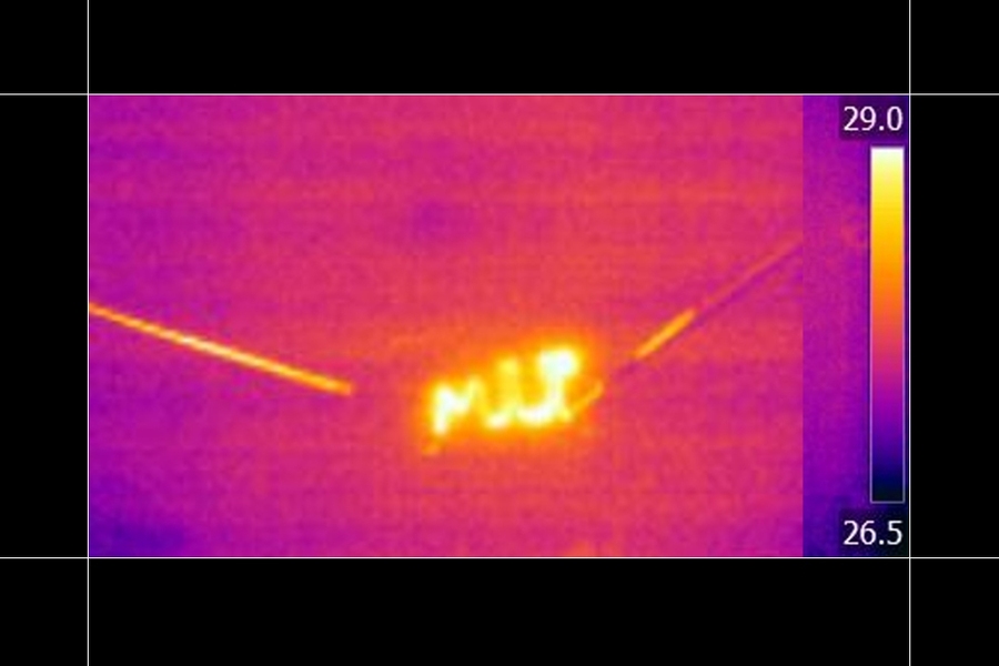 Thermal infrared camera image showing a hot spot glowing white and orange along an electrical power line against a purple-toned background, with a temperature scale ranging from 26.5 to 29 degrees Celsius