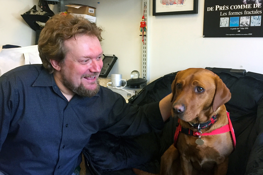 A man with curly hair and a beard smiles at a brown dog with a red harness seated beside him in a cluttered office.