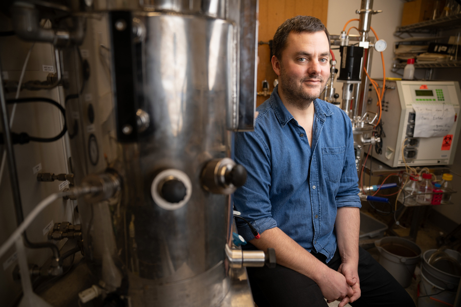 An MIT researcher in a denim shirt leans against a large stainless steel chemical synthesis vessel surrounded by lab tubing and instrumentation in a chemistry laboratory.