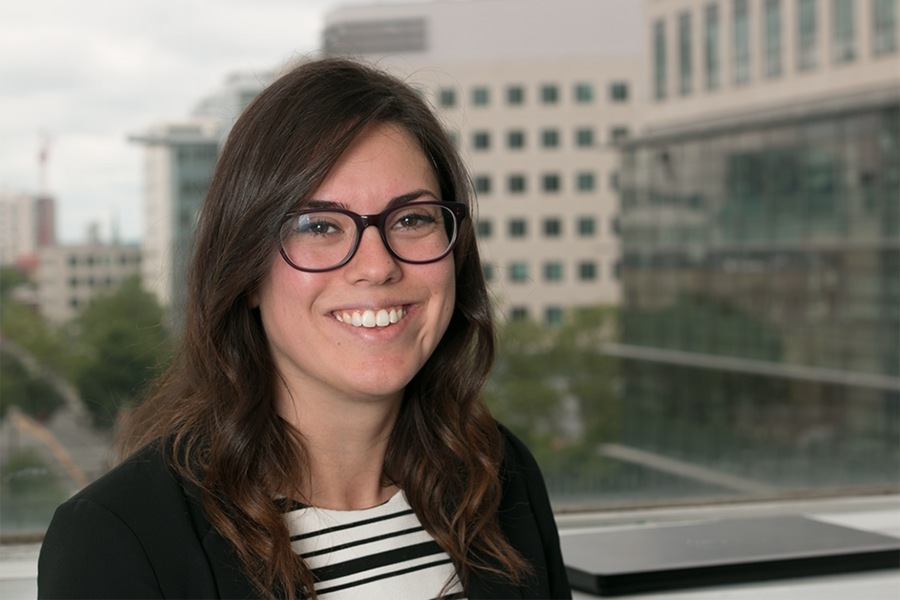 A woman with glasses and long dark hair smiles at the camera in front of a window overlooking an urban skyline.