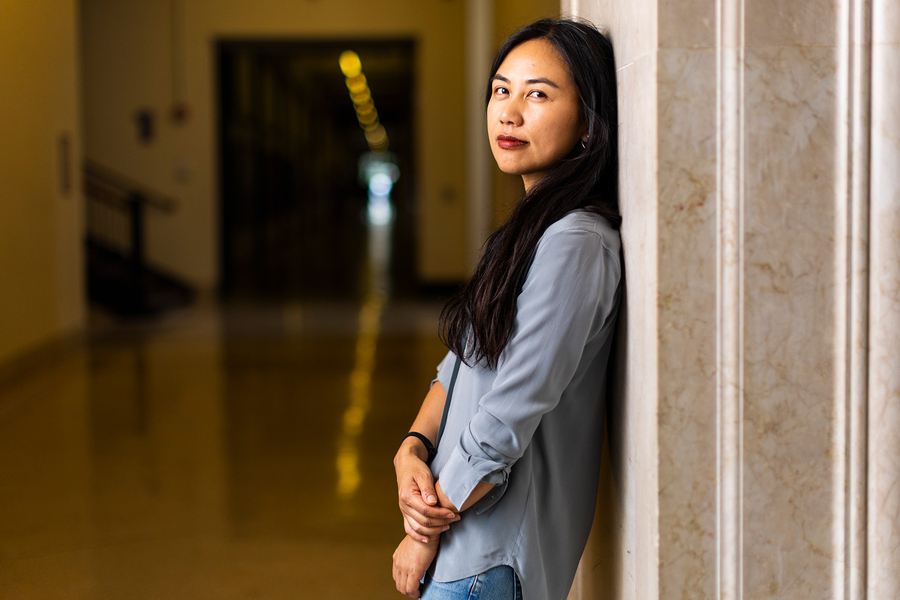 A woman in a gray blouse leans against a marble column in a grand institutional hallway, looking thoughtfully to the side.