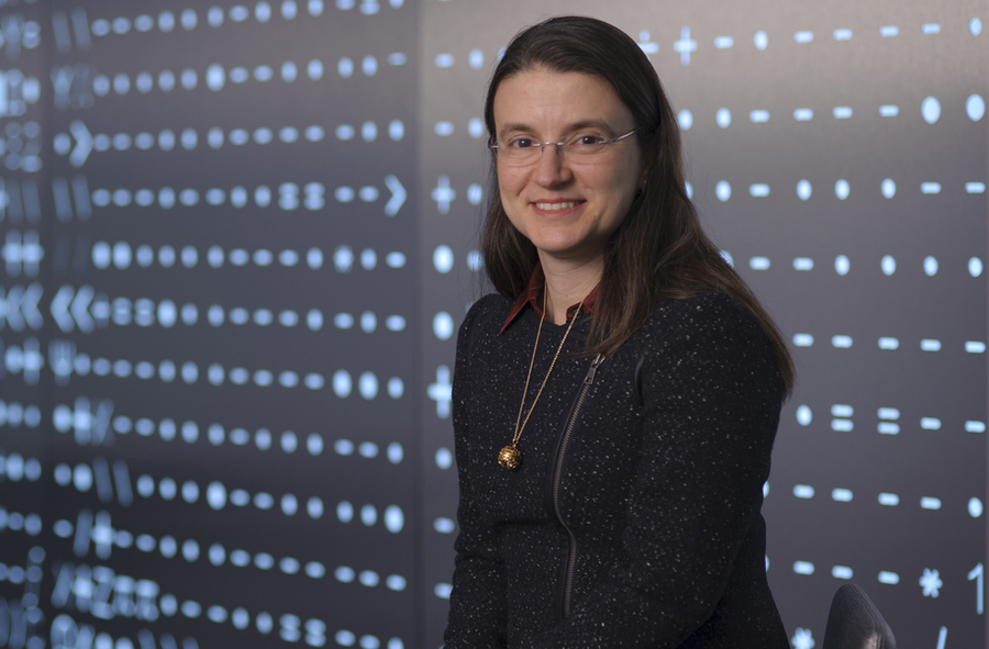 A woman with long brown hair and glasses smiles at the camera in front of a glowing blue wall covered in symbols and characters.