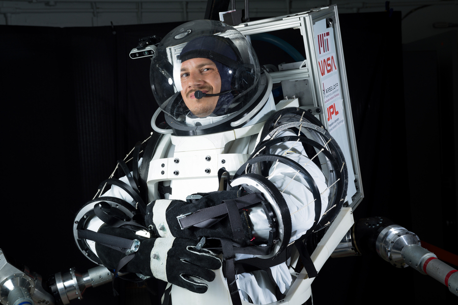 A researcher smiles while wearing a prototype MIT-NASA spacesuit with a clear helmet visor, articulated arm joints, and a backpack support frame in a dark testing facility