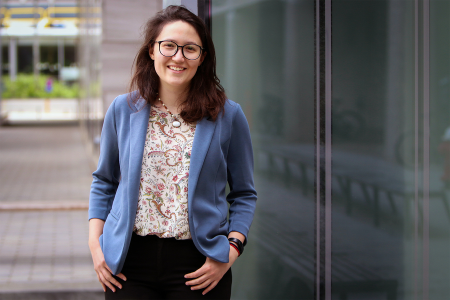 A woman in a blue blazer and floral top leans against a glass-paneled building exterior, smiling at the camera.